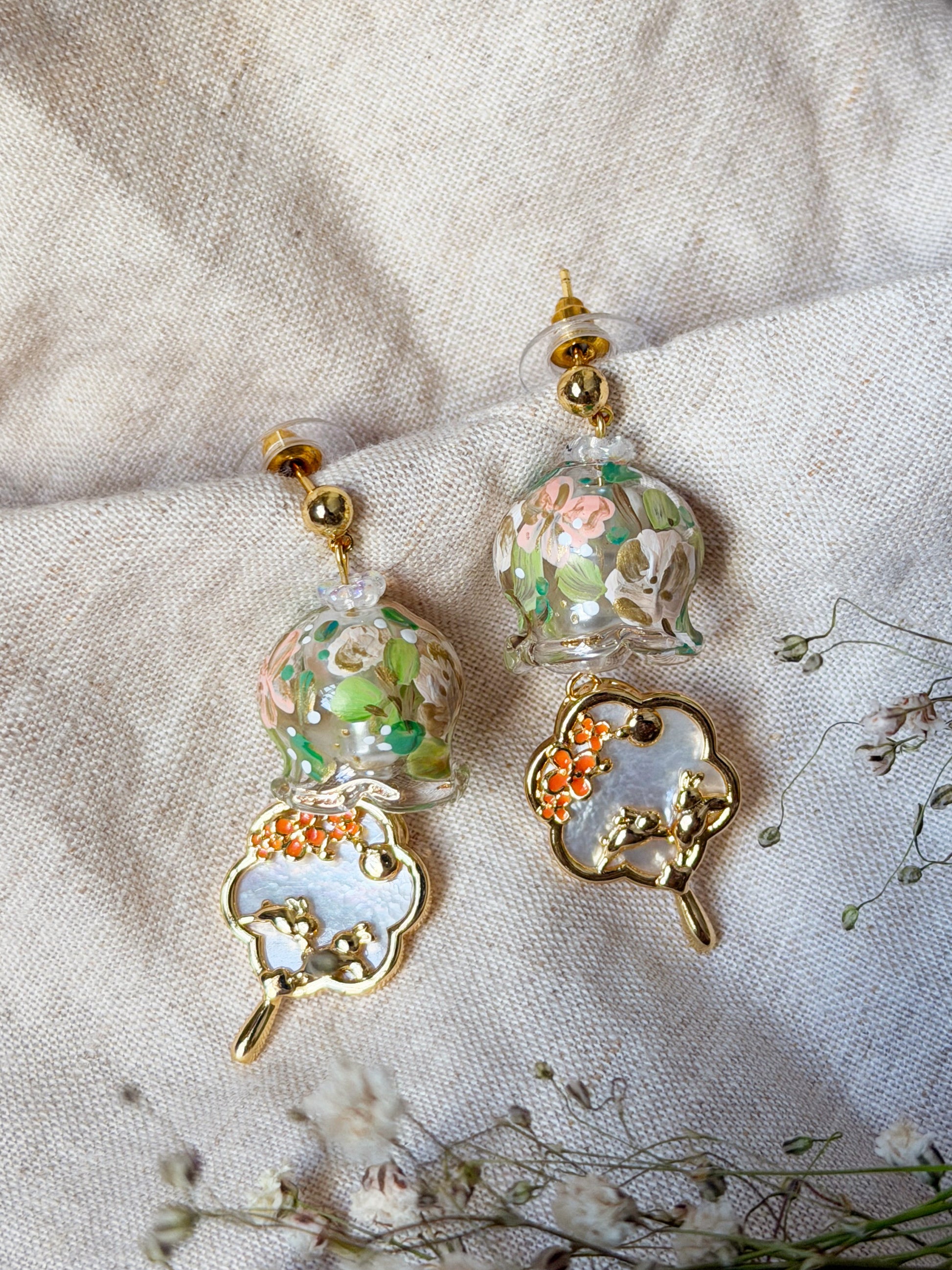 A pair of handmade gold-tone dangle earrings with colorful floral glass beads and white mother-of-pearl charms, displayed on a textured beige linen fabric with dried white flowers.